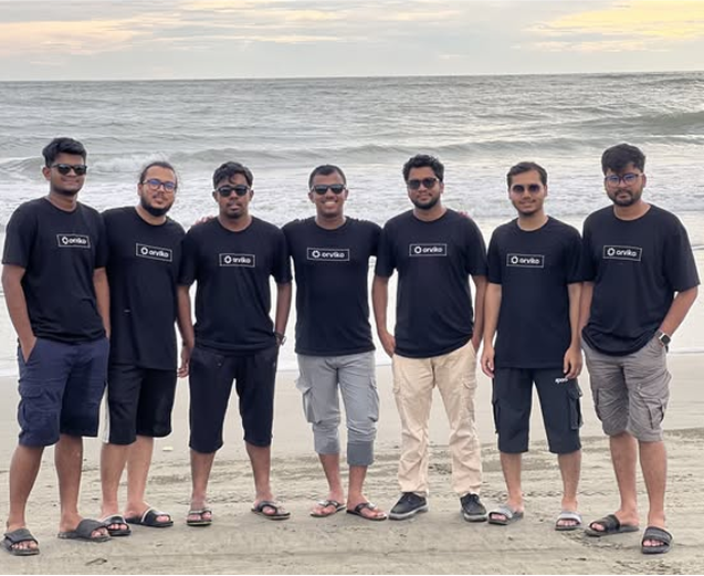 Group of seven men in matching black shirts on a beach, arms around each other, smiling.